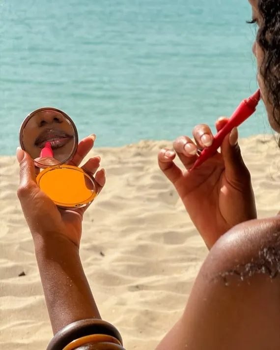 Hands holding an orange compact and a red lipstick on the sand