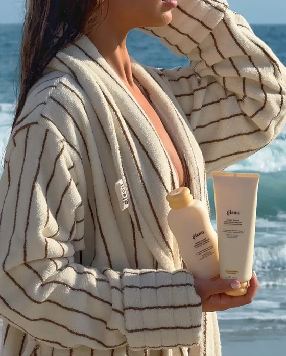 Person in a striped robe holding two skincare tubes by the sea