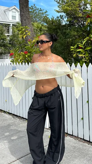 Creator in a sheer mesh top and black trousers beside a white picket fence